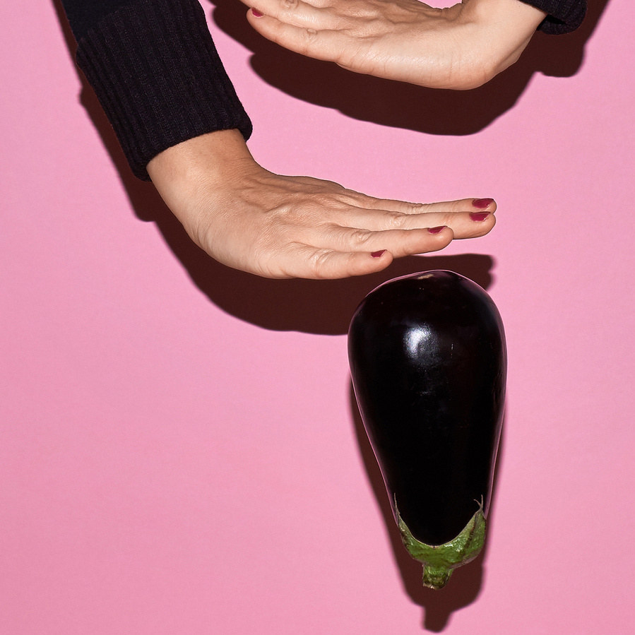 Detail of two female arms wearing a black shirt, hands with varnished finger nails playing with an aubergine in front of a plain pink background
