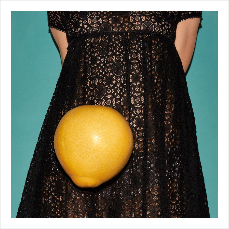 Part of a woman wearing a black lace dress with a flying yellow pomelo in front and a petrol colored background