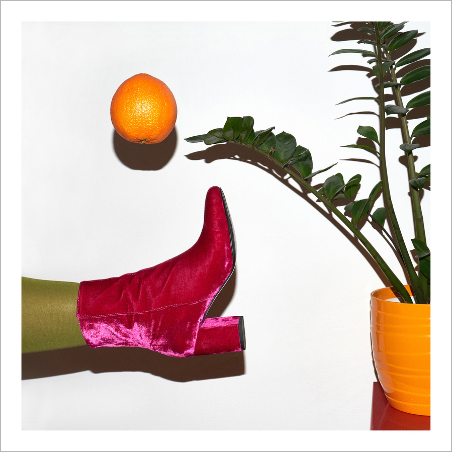 A pink woman's leg wearing a pink velvet ankle boot and green tights kicking an orange in the air in front of a white wall next to a plant in an orange flower pot on a red sideboard