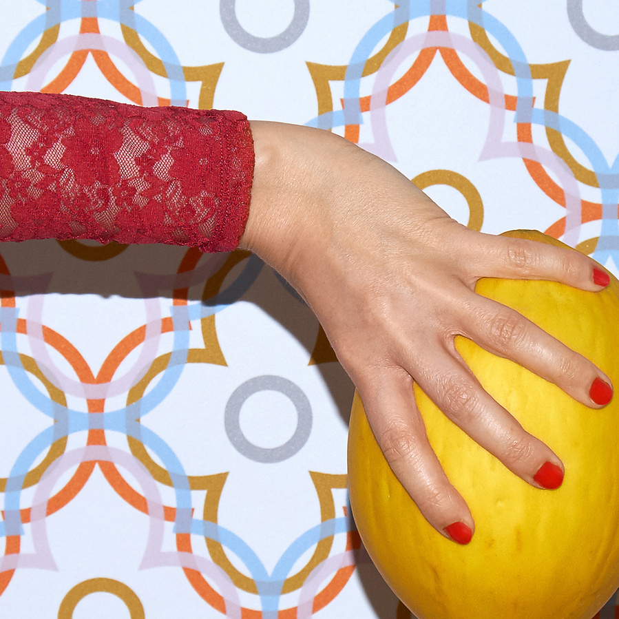 Detail of a female arm wearing a pink lace shirt, her hand with varnished red finger nails holding a yellow melon in front of a colorful patterned wall paper