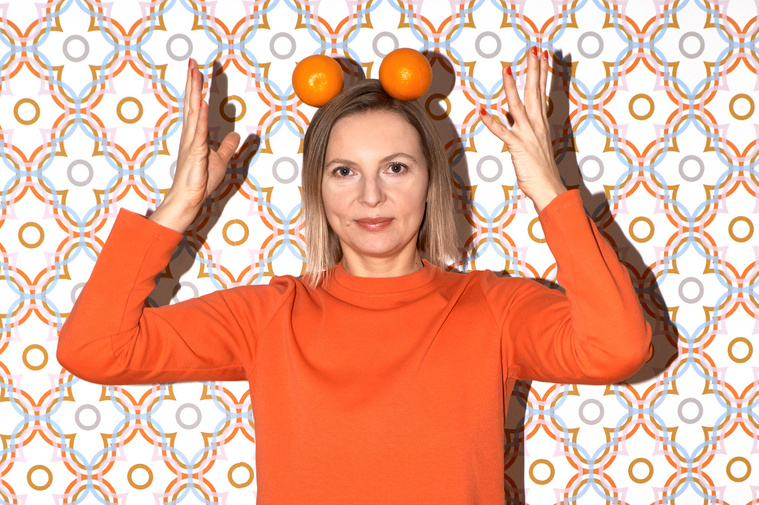 Artist N. A. Vague wearing an orange dress, throwing two oranges above her head, in front of a colourful patterned wallpaper