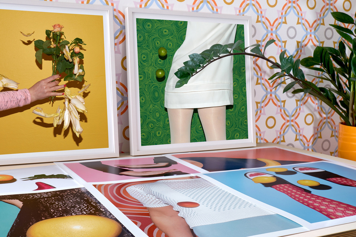 Colorful art prints by N. A. Vague on a desk in front of a colorful patterned wall paper, next to a plant in an orange flowerpot