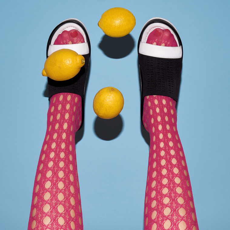 Two female legs wearing pink tights and black and white shoes behind three flying lemons in front of a plain light blue background