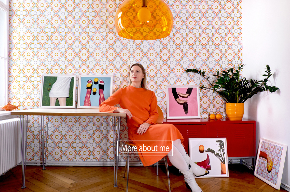 Artist N. A. Vague wearing an orange dress and white tights sitting on an orange chair with framed prints of her artworks in front of a colorful patterned wallpaper