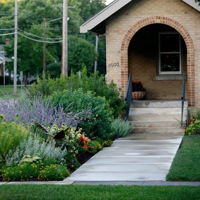A lush, naturalistic front yard planting with Perovskia, Echinacea, Amsonia, and Musa along a welcoming entry path to a brick home.