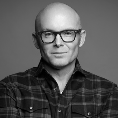 Black and white portrait of Adam Woodruff, a garden and landscape designer, wearing a plaid shirt and glasses. He has a confident expression against a neutral background.