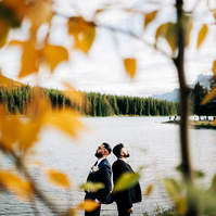 A same sex couple standing back to back with a lake behind them.
