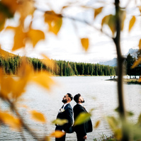 A same sex couple standing back to back with a lake behind them.