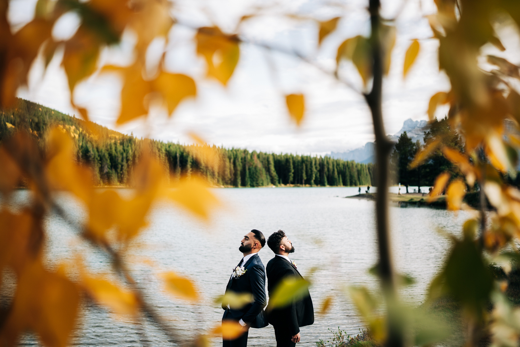 A same sex couple standing back to back with a lake behind them.