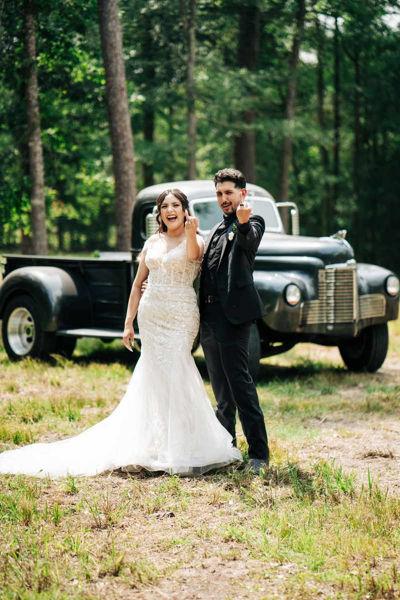 A newlywed couple flicking off the camera with a classic truck behind them.