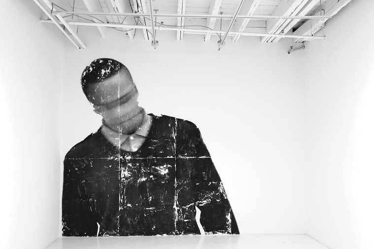 Photograph of a grey-toned projection that depicts an antique image (or altered to appear that way)  of a person of colour (their face is predominantly covered by a fedora) with a shatter texture overlaid; in a dark gallery.