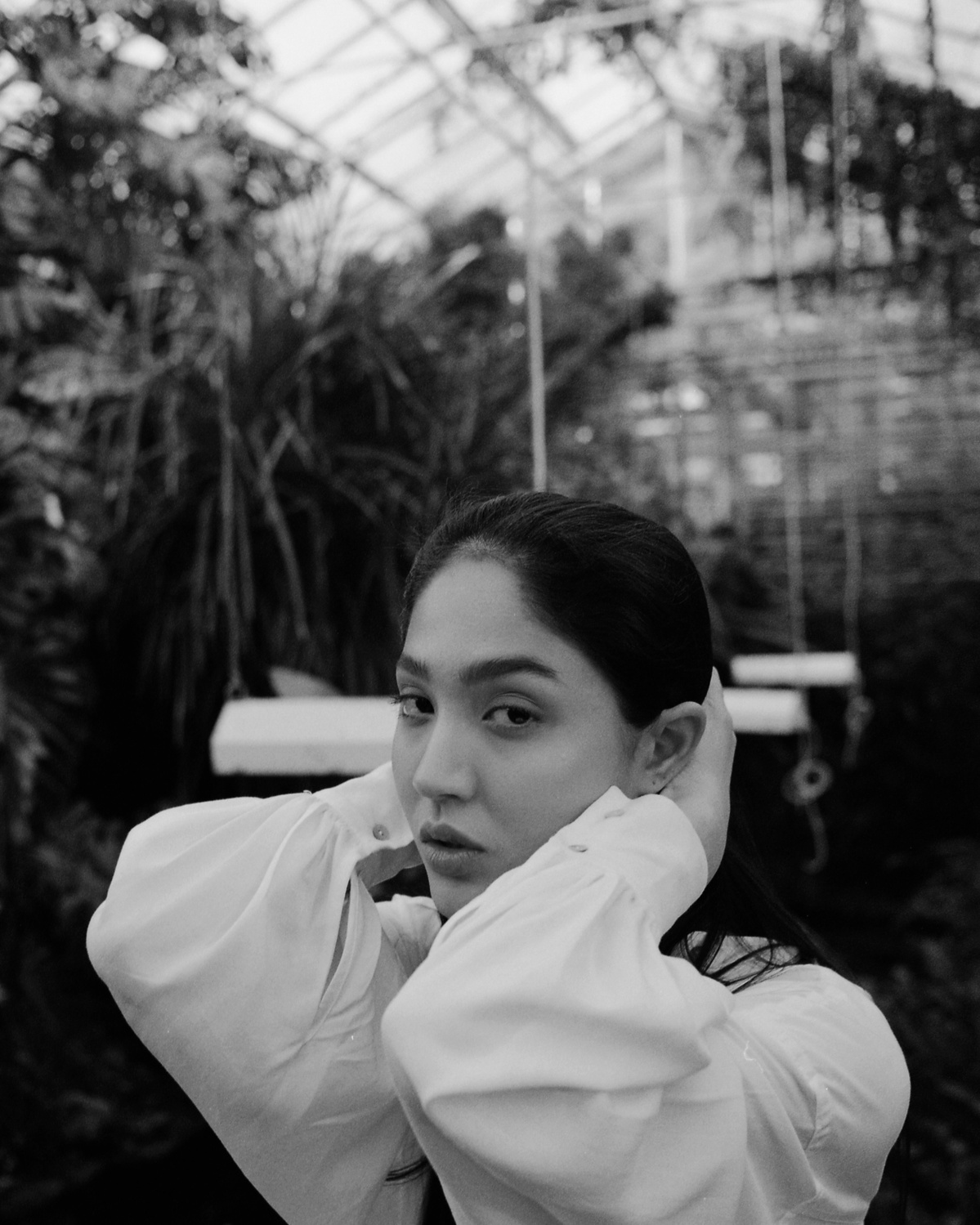Portrait of an Asian woman in the greenhouse. She is holding her neck and wears a white, wide blouse.