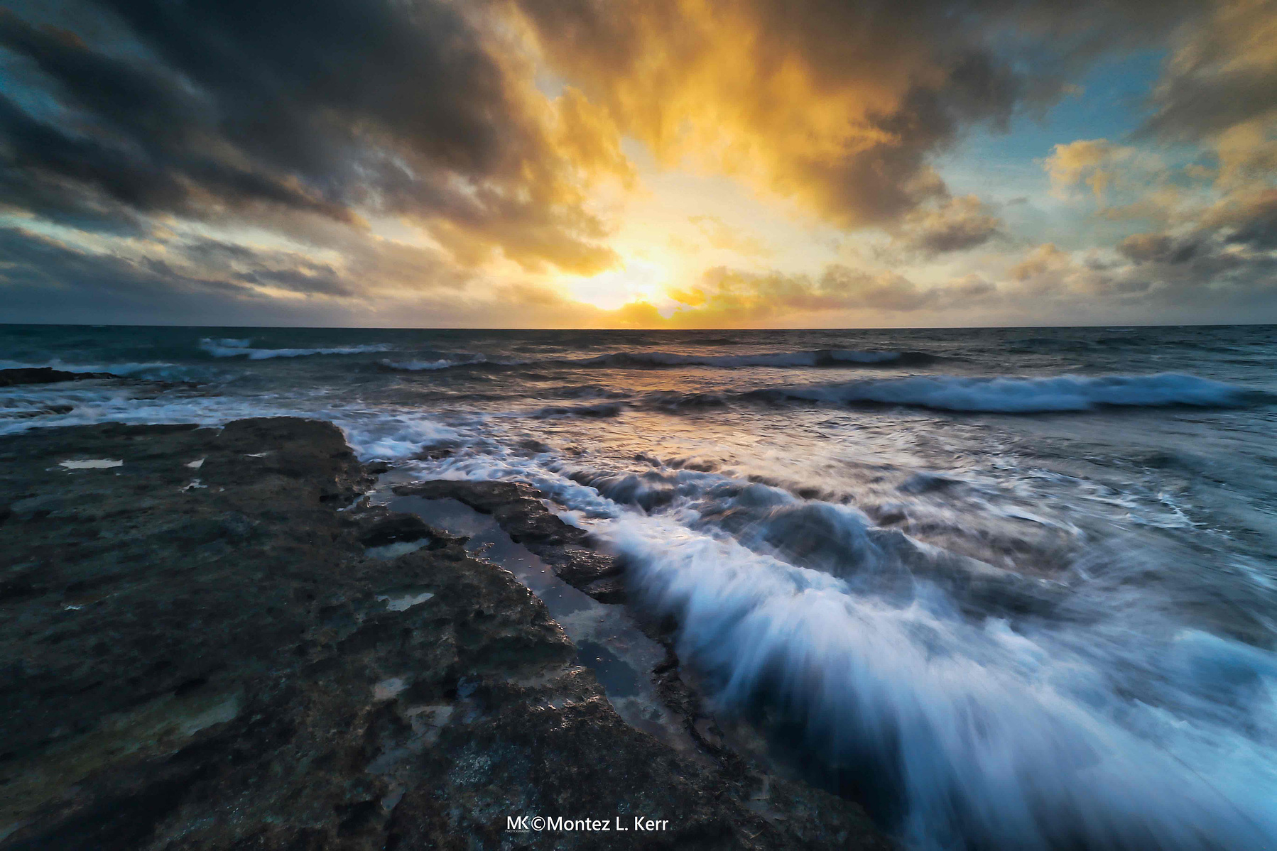 Waves crash against a rocky shoreline under a vibrant sunset sky, casting a warm glow on the water. Dark clouds add drama and contrast to the colorful horizon.