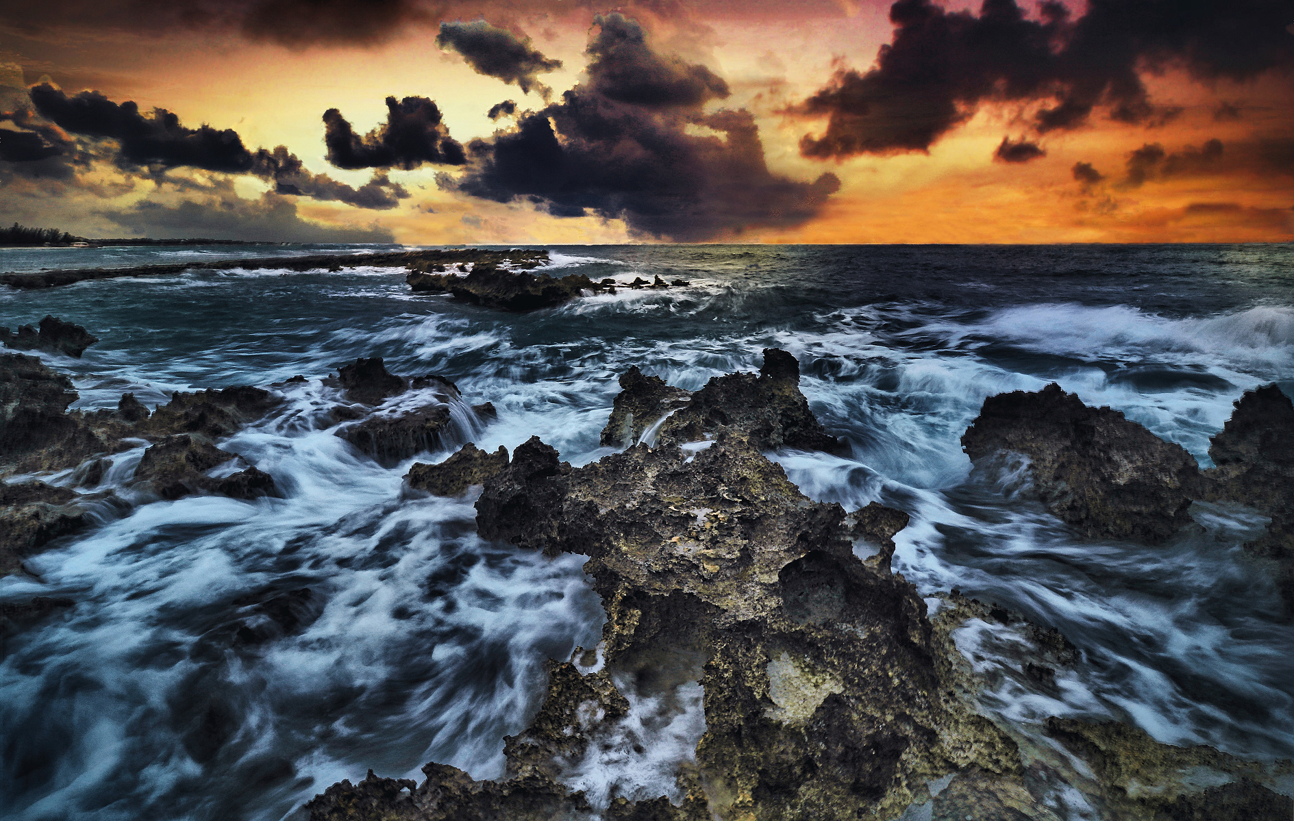 Dark, dramatic clouds loom over a turbulent sea, with waves crashing against jagged rocks. The vibrant sunset casts an orange and purple hue across the sky, creating a stunning contrast.