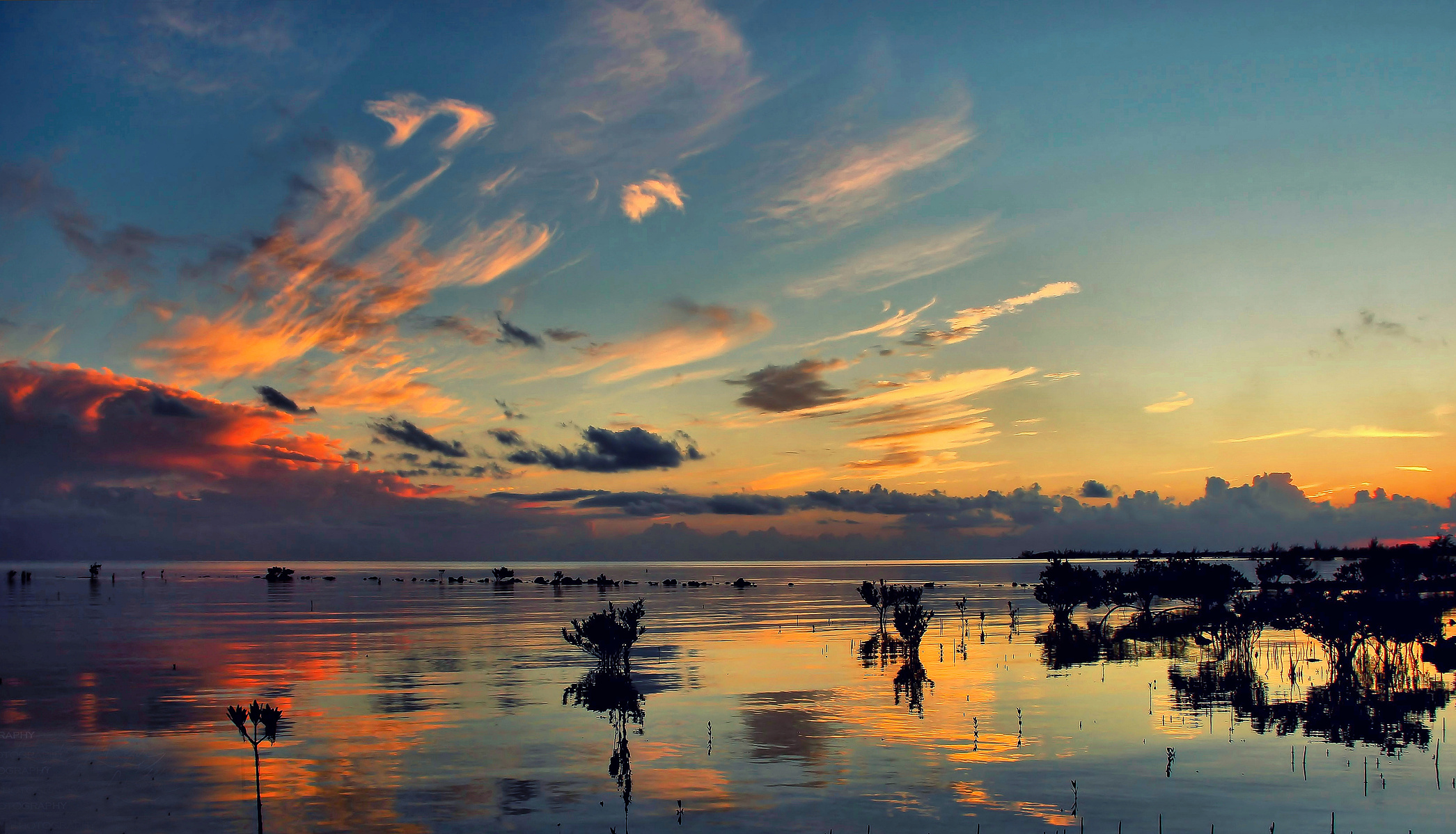 A breathtaking sunset over a tranquil body of water creates stunning reflections of the colorful sky. Wispy clouds stretch across the scene, adding dynamic contrast against the fading daylight.