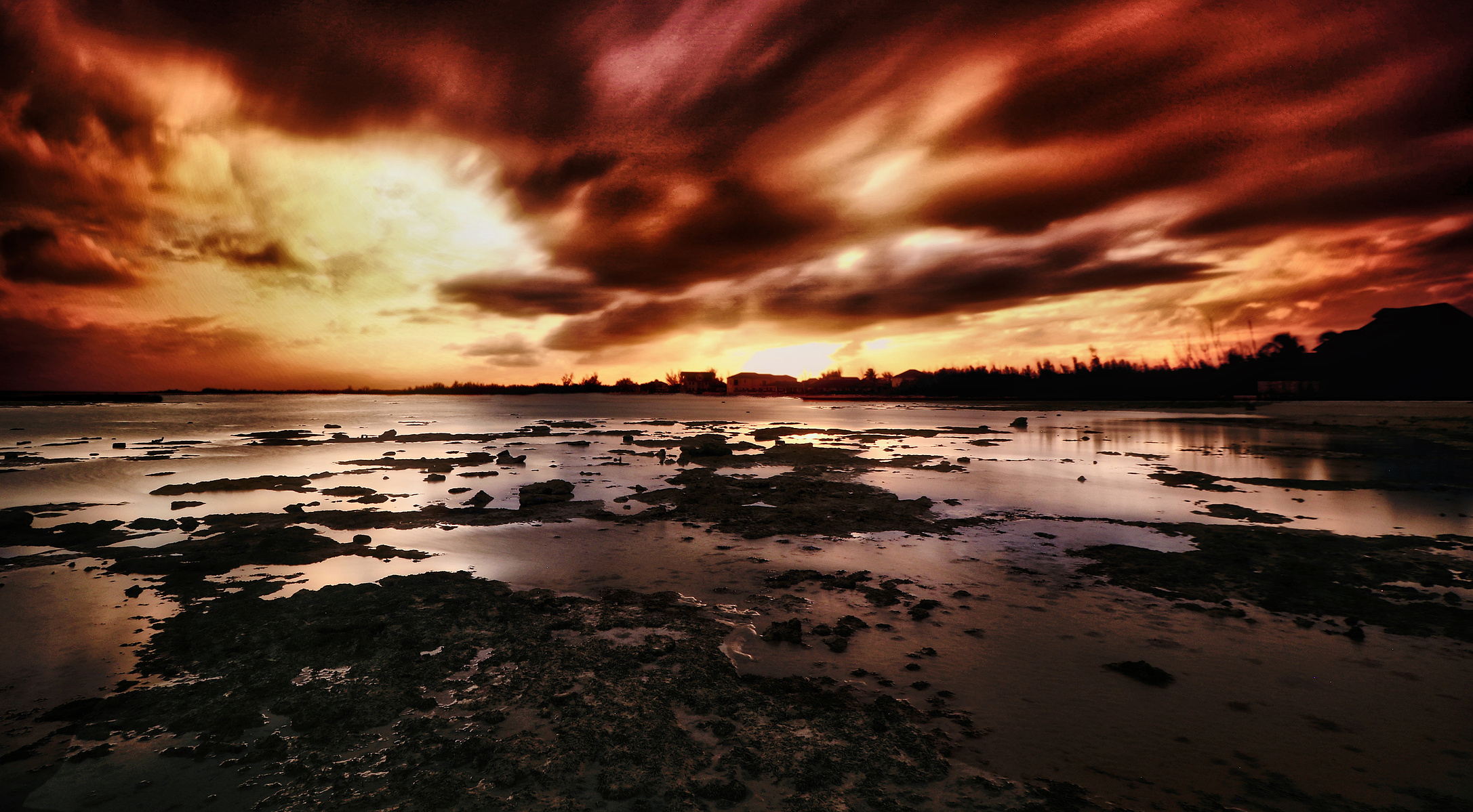A dramatic sunset casts vibrant colors across the sky, reflecting off a calm water surface scattered with rocky outcrops. The silhouette of a distant tree line adds contrast to the luminous horizon.