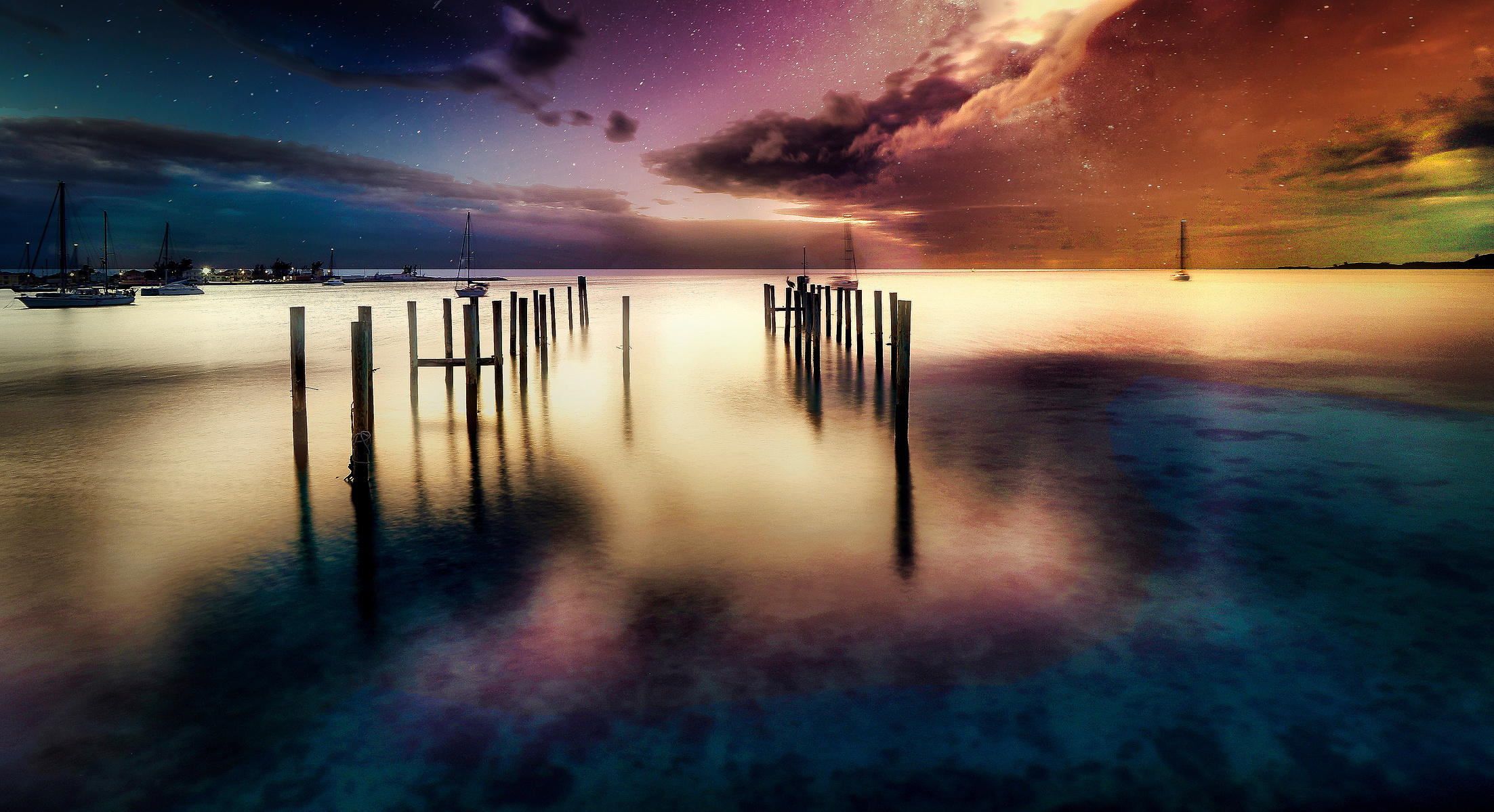 A tranquil seascape features a series of wooden posts emerging from calm waters under a sky transitioning from deep blue to warm orange hues. Sailboats are faintly visible on the horizon, and the water reflects the vibrant colors of the sunset and night s