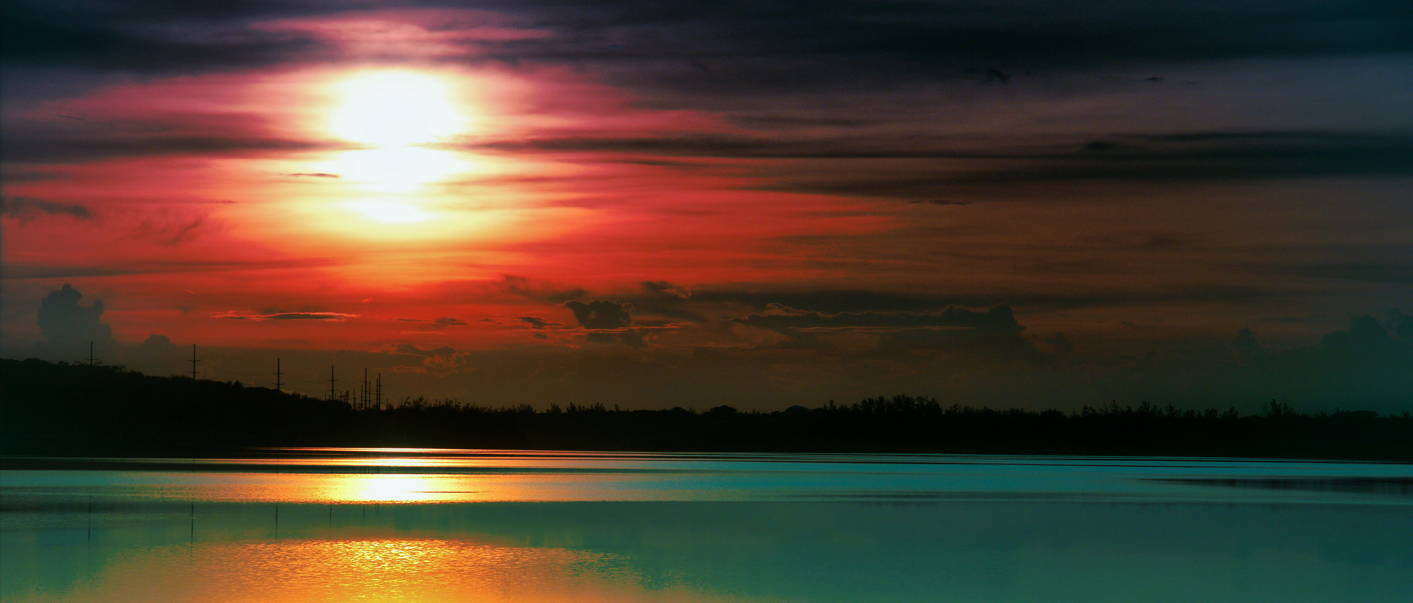 A tranquil sunset scene unfolds over a calm body of water, with a vibrant red and orange sky reflecting on the surface. Silhouettes of distant trees and clouds add depth to the serene landscape.