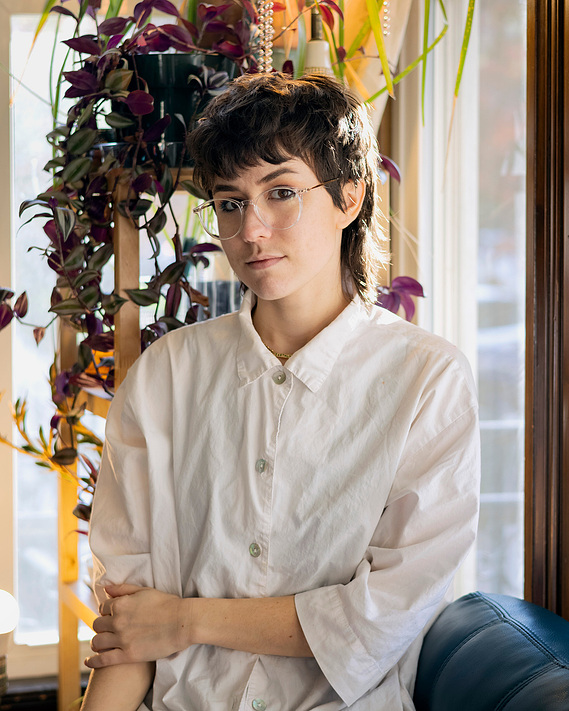 A young white woman with a pixie haircut and glasses, sitting in front of a window while looking calmly into the camera. 