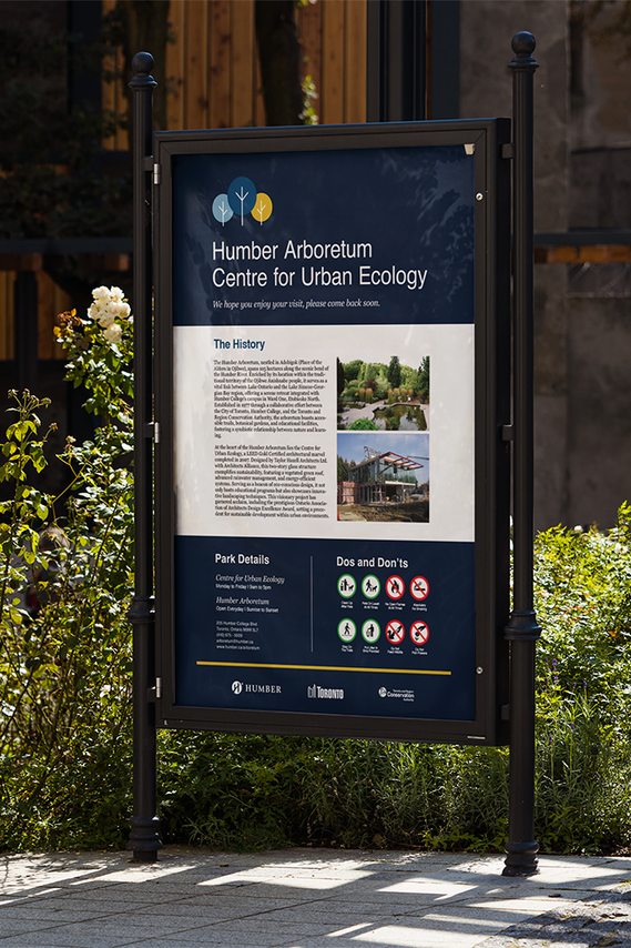 The image displays a large sign on the park pathway, showcasing Humber Arboretum's history, information, and rules. White text contrasts against a dark blue background, with construction images included.