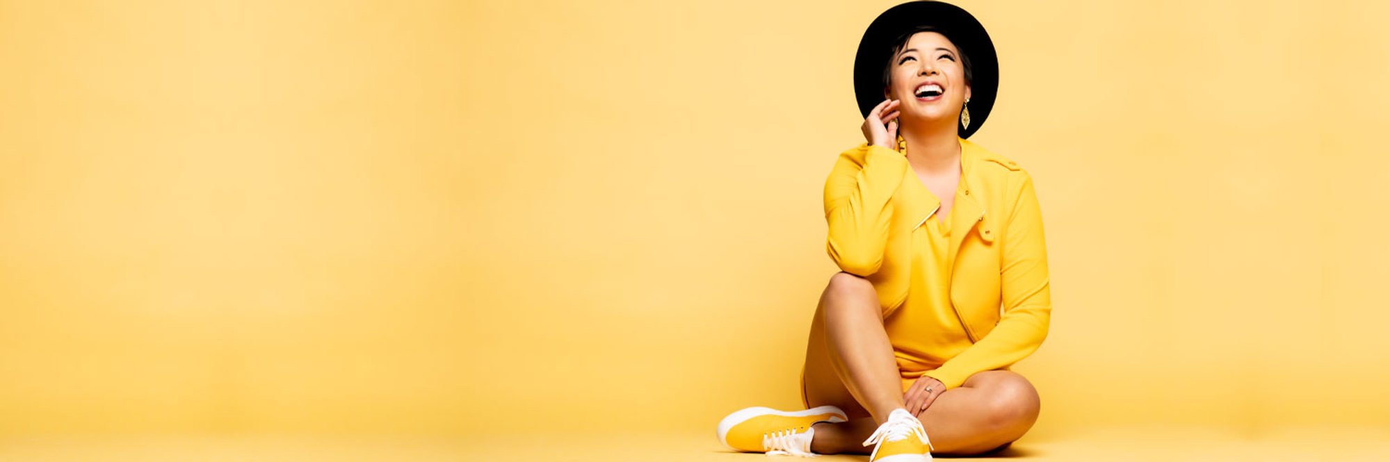An Asian woman wearing a yellow jacket, yellow shirt, yellow shorts, and yellow shoes and a black hat sits on a yellow background and laughs uptowards the ceiling