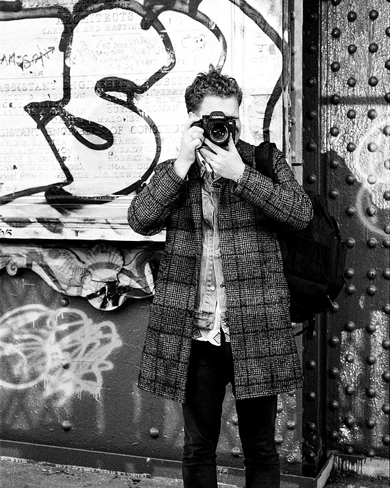 The image is of Drew Chambers. It is in black and white. He is taking a photo with his camera up to his face. He is wearing black pants and a patterned overcoat. He has curly hair.