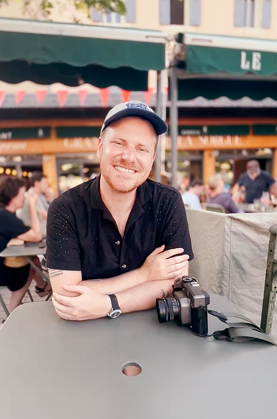 The image is of Drew Chambers. He is white, has short facial hair, and is wearing a baseball cap, a short sleeve button shirt, and a watch. He is sitting outside at a cafe. A Mamiya 7 film camera is in front of him on the table. 