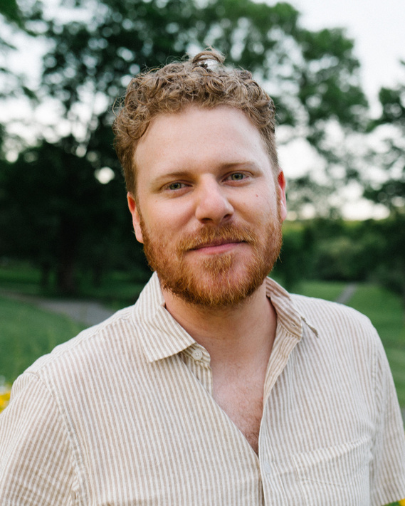A photo of Drew who has red, curly hair, a beard, and green eyes. He is wearing a light taupe and white striped button-up shirt. Behind him is some foliage of green grass, trees, and white and yellow flowers. He is softly smiling and looking at the camera