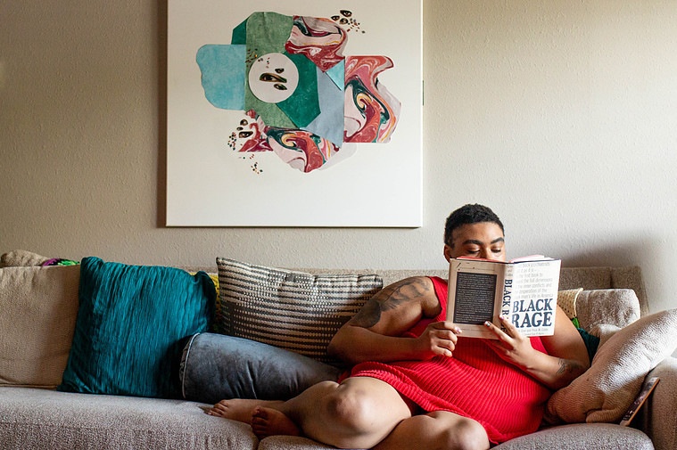 A  Black fat person is wearing a red dress and holding a book that says Black Rage. The person Is sitting on a couch with a bunch of pillows