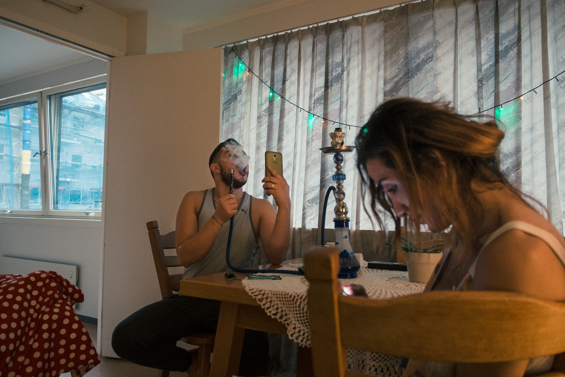 Omar (left) smokes a nargila and chats with his mother via video in Mary's (right) apartment. The two are both Syrians living in the same apartment block in Bergen, Norway.