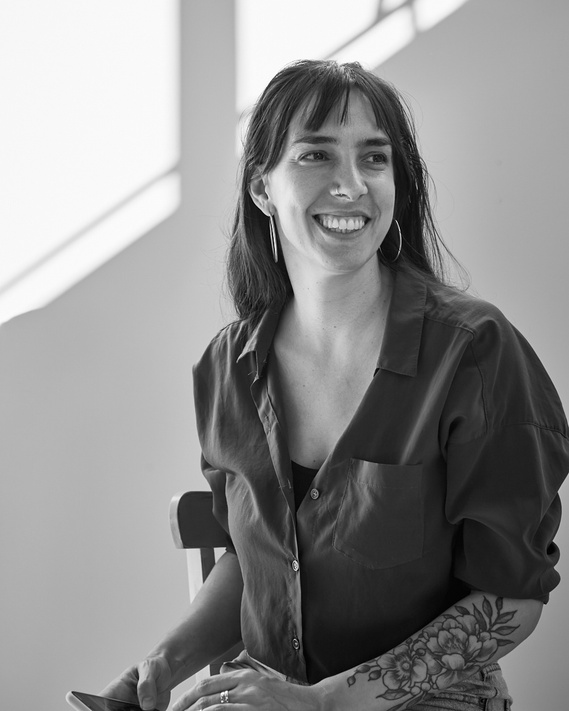 Black and white portrait of Magali Polverino. She is looking at her left with a smile on her face. She has a dark shirt and a flower tatoo on her arm.