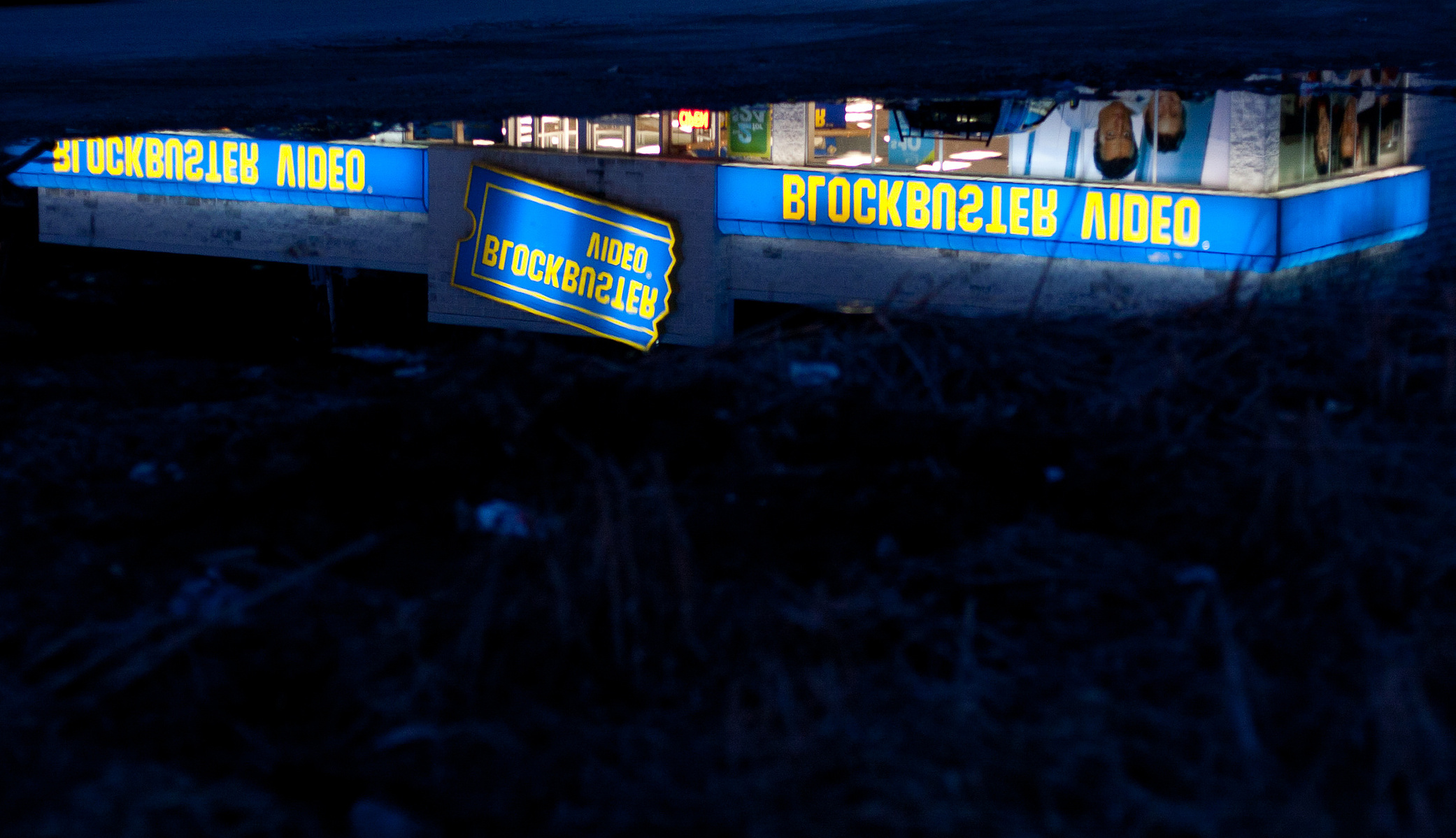 Illuminated blockbuster video sign reflected in puddle.