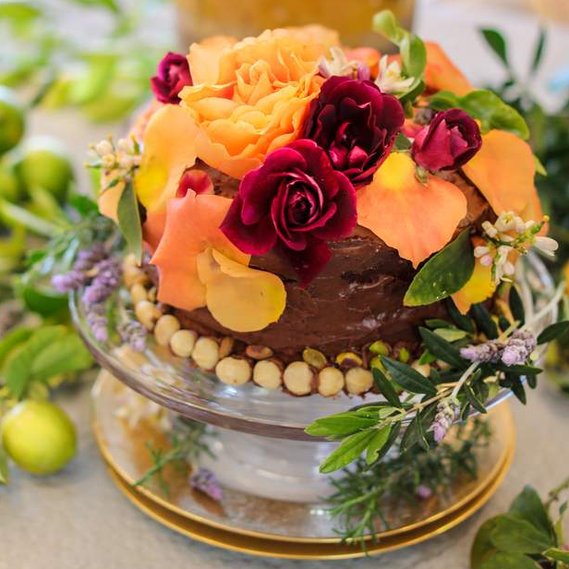 A chocolate olive oil cake decorated with pistachios and macadamia nuts. Covered in vibrant roses. 
