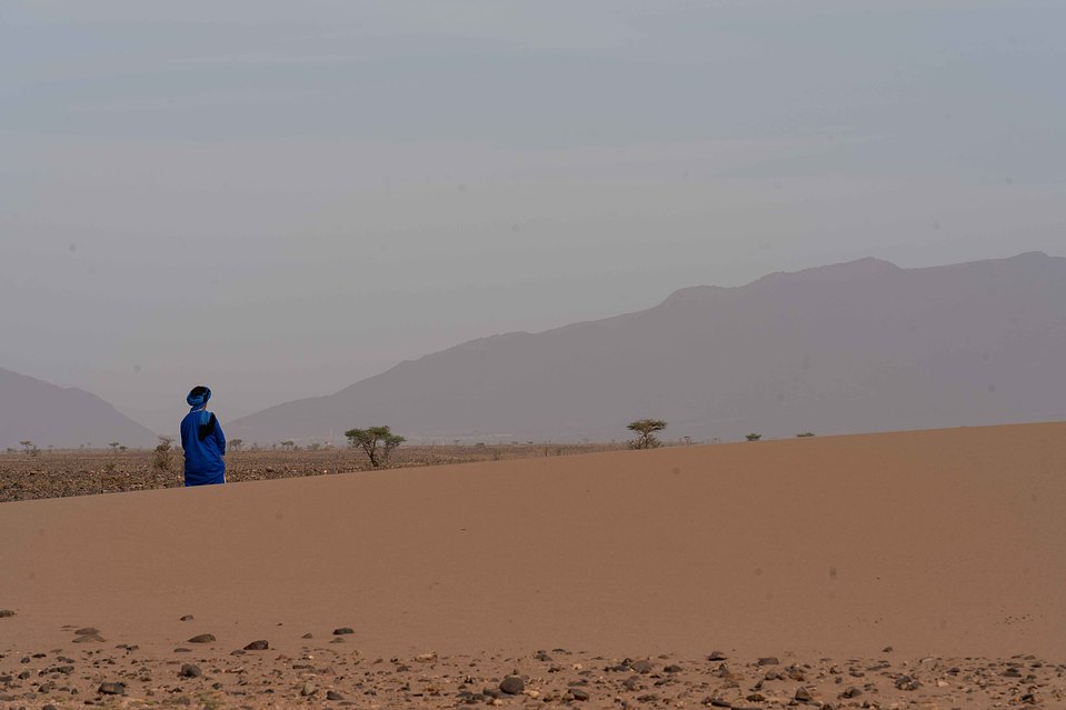 Nomade vêtu de bleu dans le désert du Sahara au Maroc, capturé sous une lumière douce avec les montagnes en arrière-plan.