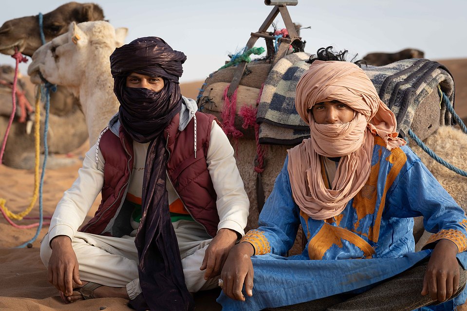 Deux hommes nomades posent fièrement aux côtés de leurs dromadaires dans le désert du Sahara, incarnant la résilience et la connexion profonde avec cet environnement aride et majestueux.