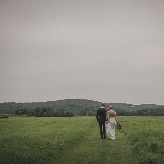 Couple walking hand in hand away from the camera beneath a bright, summer overcast sky at Lion Rock Farm, surrounded by lush green mountains, captured by a Connecticut wedding photographer.