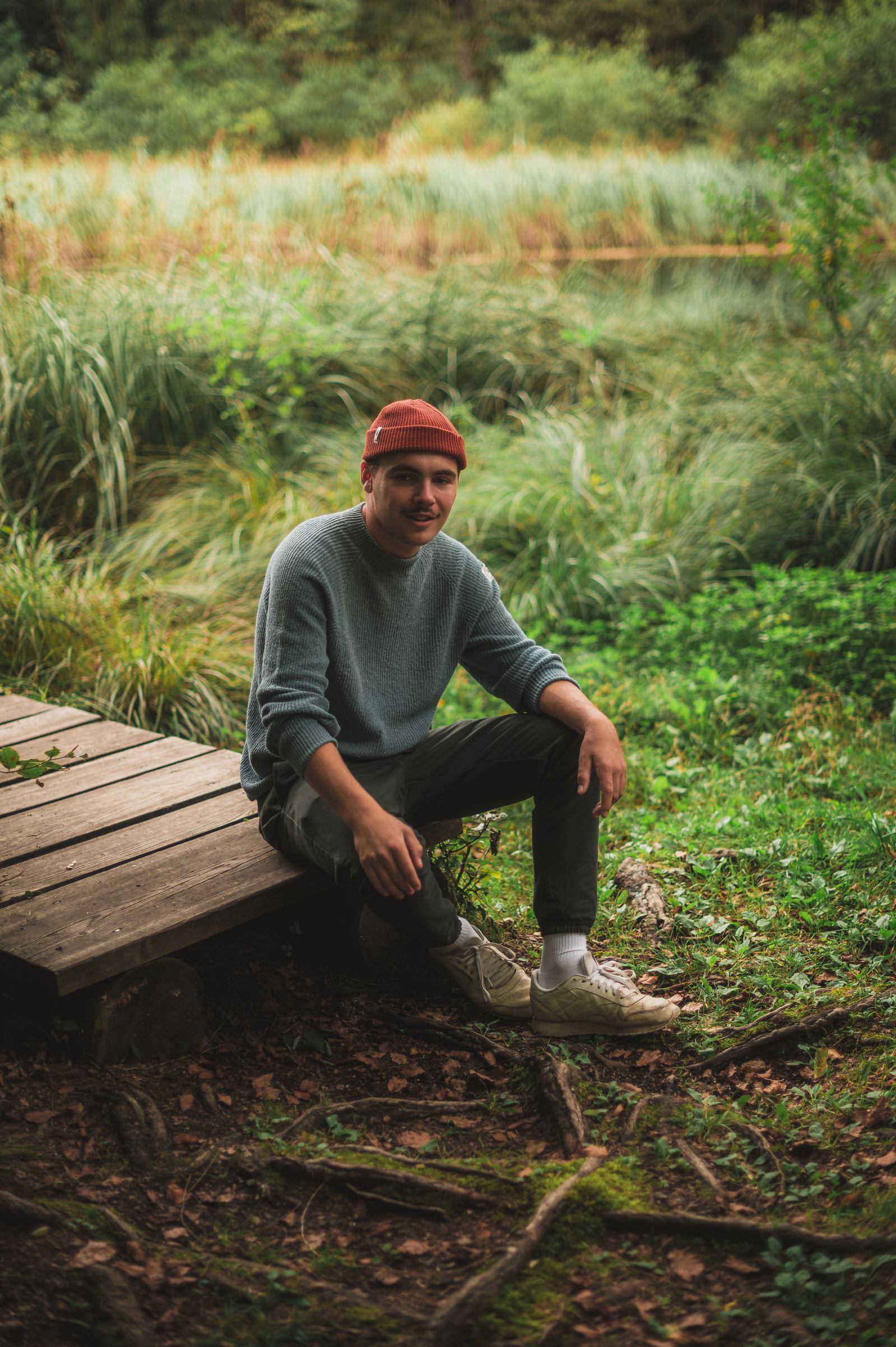 Actionshoot Shot: Mann mit Mütze trägt Pullover, sitzt auf kleiner Holzbrücke im Wald.