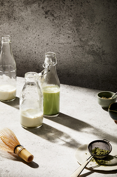 Matcha latte drink in clear milk bottles on a light rustic slab with a grey textured background aesthetically styled by a food stylist and photographed by a professional food and drink photographer