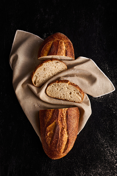 Fresh baked loaf of sourdough baked and sliced on a beige rustic linen cloth sitting on a dark bench with some flour spilt to the side