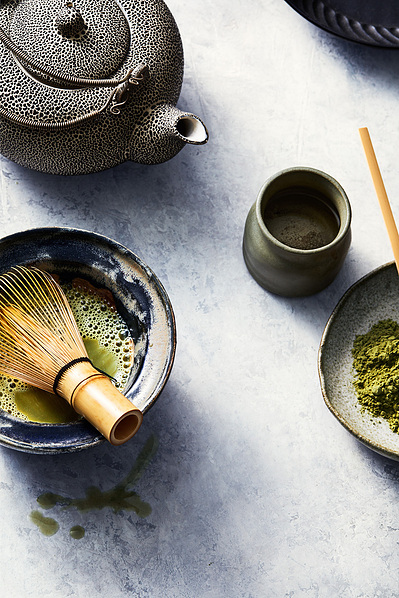 Matcha latte drink in a rustic mixing bowl with a matcha whisk with a kettle and matcha powder on the sides, sitting on greyish blue textured background aesthetically styled as an editorial and photographer by professional drink photographer