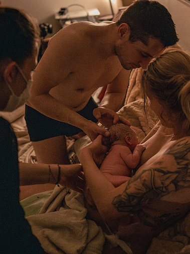 Le nouveau né est dans les bras de sa maman pour la première tétée, le papa aide le bébé et la sage femme veille à ce que tout se passe bien.