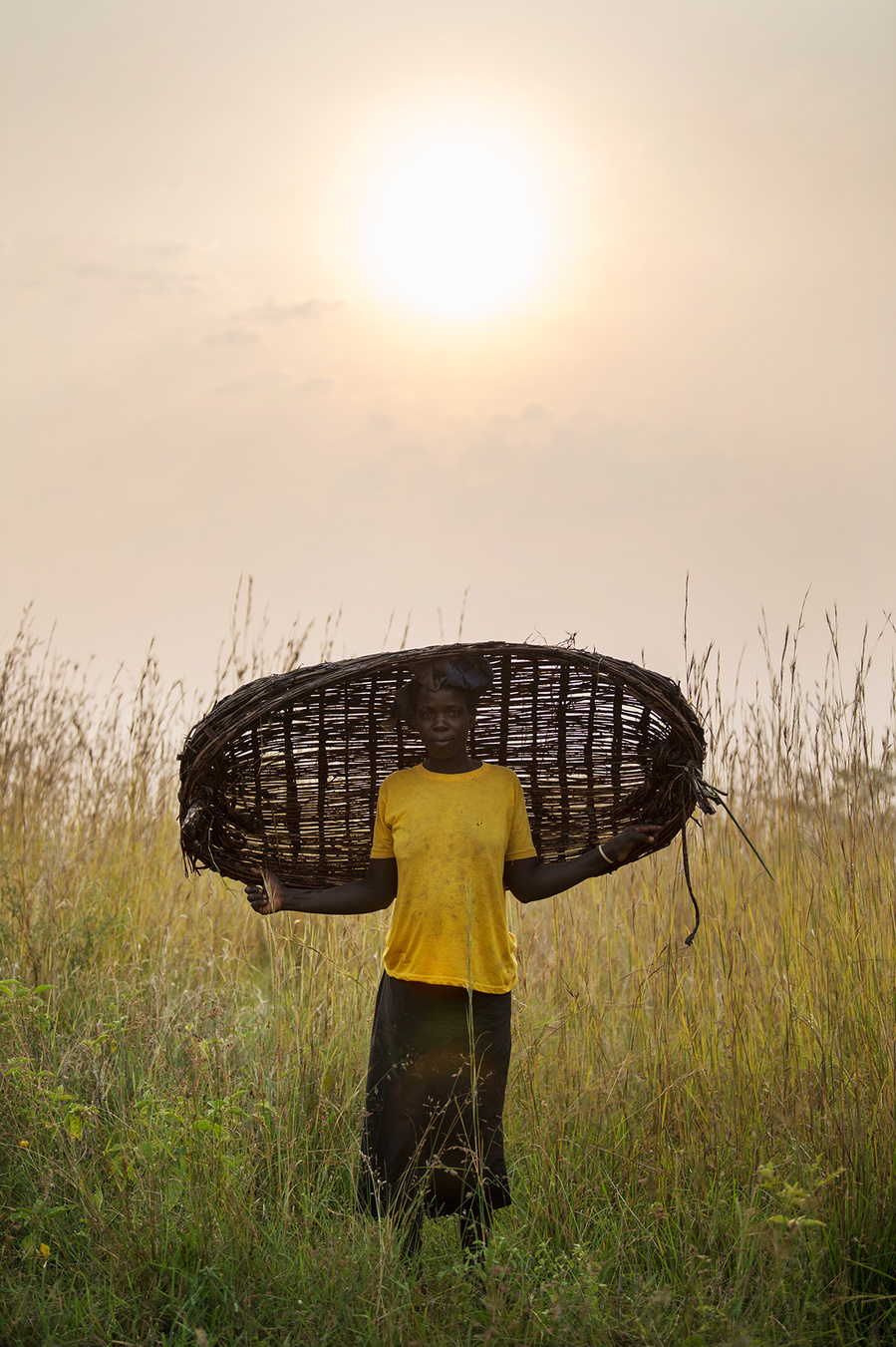 A fine art print of a woman walking through the grass with her fishing basket at sunset in Uganda, Africa.
