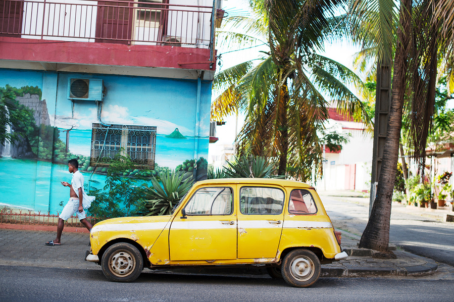 A man passes a yellow R4 Renault parked next to a palm tree, and in front of a building with beach painted on the wall in Madagascar. 