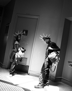Two African American children, a boy and a girl, that are tourists in Ellis Island are waiting outside of a bathroom. They are both wearing Statue of Liberty crowns.  