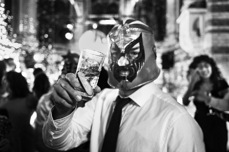 A man in a luchador mask holding up a drink in the middle of a wedding reception in London England
