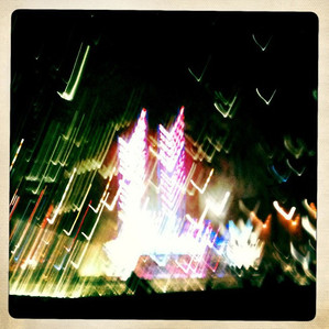 A night time photo of a person sitting at a fountain as the lights of Radio City Music Hall are streaking.