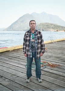 A man standing in a rigid pose with a stoic look on a dock that looks across a bay to steep mountains.