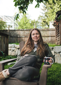 A morning coffee is enjoyed by a young woman in a garden showing lush spring plant life.
