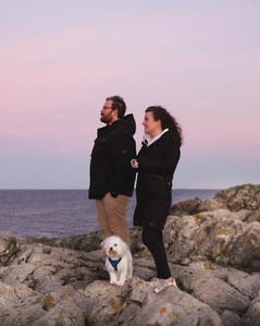 A couple in black coats look for whales across the strait while their small white dog inspects the photographer.