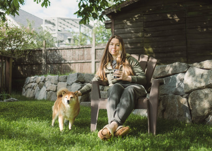A young woman reaches out to pet her dog while they enjoy morning sun under a large maple tree.